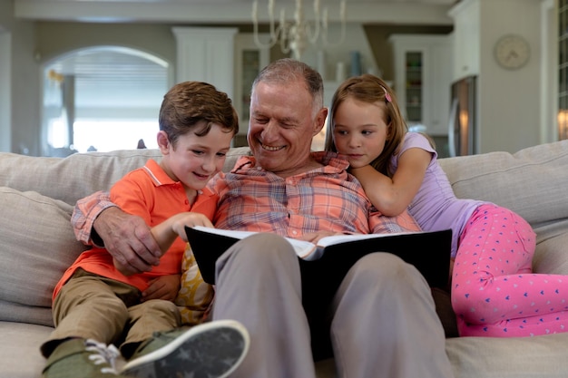 Photo low angle front view of a senior caucasian man at home in the living room sitting on the couch with his grandson and granddaughter, reading a book together and smiling