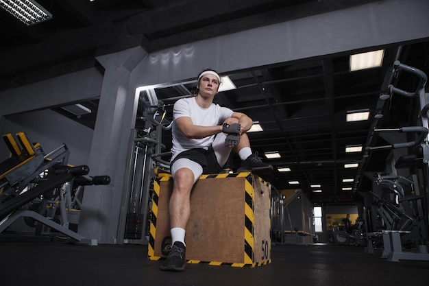 Colpo a figura intera ad angolo basso di un bell'uomo atletico seduto su una scatola di salto in palestra che riposa dopo l'esercizio