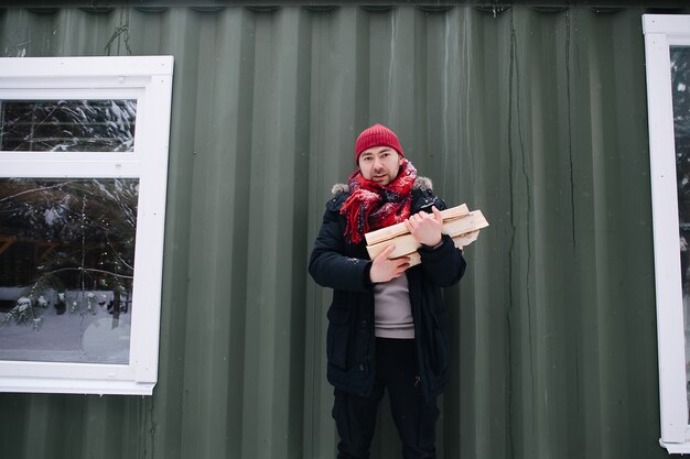 Low angle image of a man in snowy winter clothes holding logs in hands. He wears red knitted hat and scarf. Standing in front of a house wall.