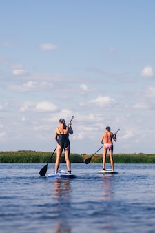 水着​を​着て​背景​に​緑​の​葦​の​波紋​の​ある​青い​湖​で​同じ​ように​オール​を​動かす​sup​ボード上​の​低​角度​の​年配​の​女性​高齢者​の​ため​の​アクティブな​ライフスタイル