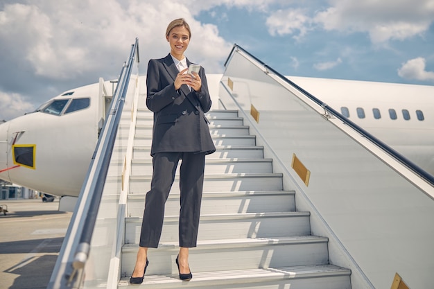 航空機の階段にガジェットを持って立っている満足している金髪の女性のローアングル