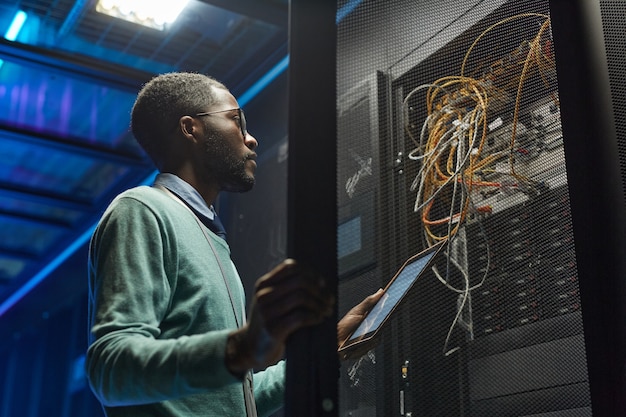 Low angle portrait of African American data engineer holding digital tablet while working with supercomputer in server room lit by blue light, copy space