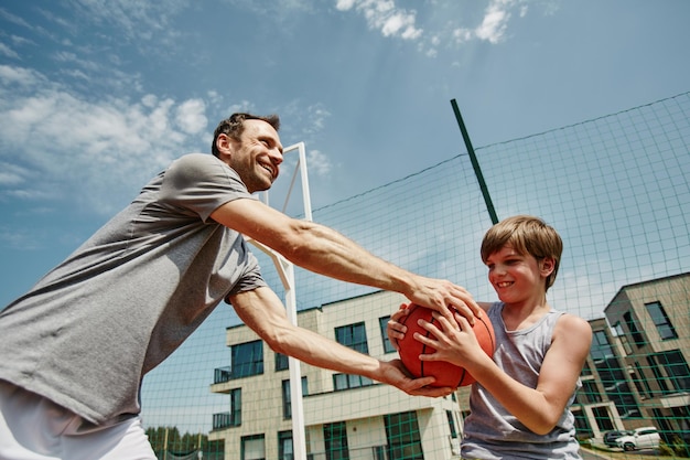 Low angle portrait of father and son playing basketball together and smiling happily against sky