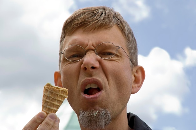 Foto ritratto a basso angolo di un uomo maturo che fa la faccia mentre mangia un cono di gelato contro il cielo