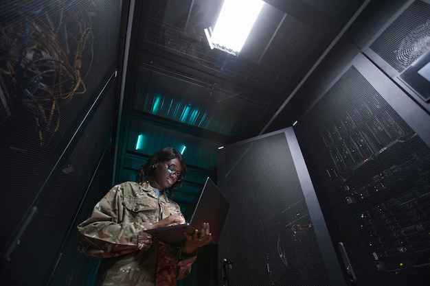 Low angle portrait of young African-American woman wearing military uniform using laptop while standing in server room, copy space