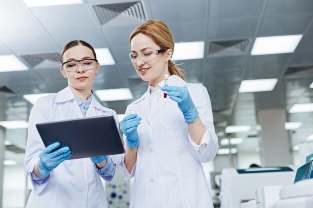 Low angle. Pretty young scientists keeping smile on faces standing in the lab while going to examine blood