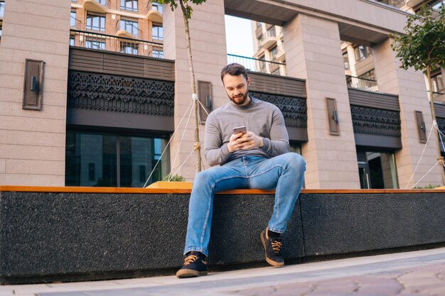 Low-angle shot of bearded young man wearing casual clothes using mobile phone while sitting on urban bench at empty European city street.