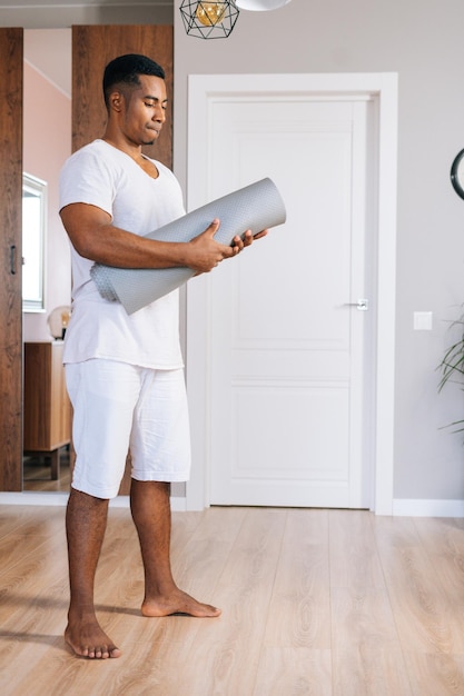 Low-angle shot of muscular African-American sportsman rolling yoga mat after finishing working out exercising at bright domestic room. Concept of sport training at home gym.