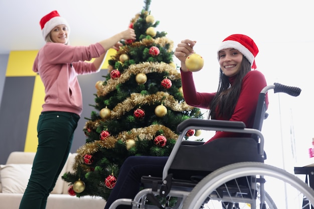 크리스마스 트리를 화려한 공, 휠체어를 탄 여성, 축제 분위기, 새해를 축하하는 자매들의 낮은 각도. 새해, 크리스마스, 장애인, 가족 개념