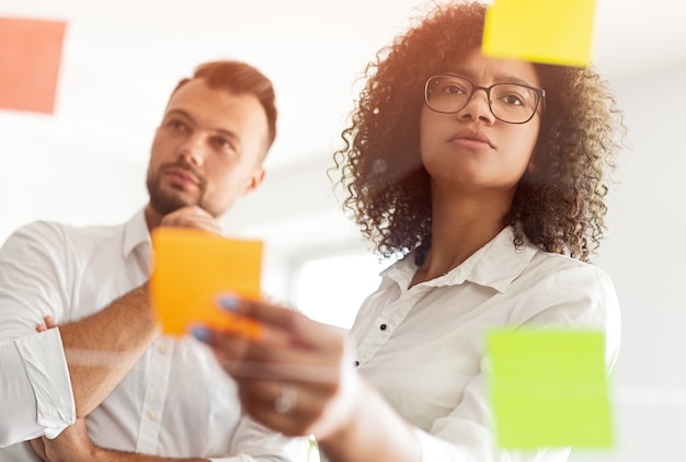 Low angle of smart black woman reading sticky notes and thinking while working on project with pensive male colleague during meeting in modern office