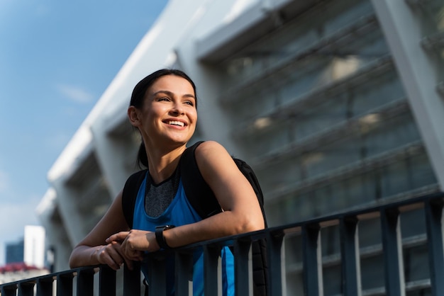 Low angle of smiling cheerful fit young woman leaned her elbows on metal handrail looking away