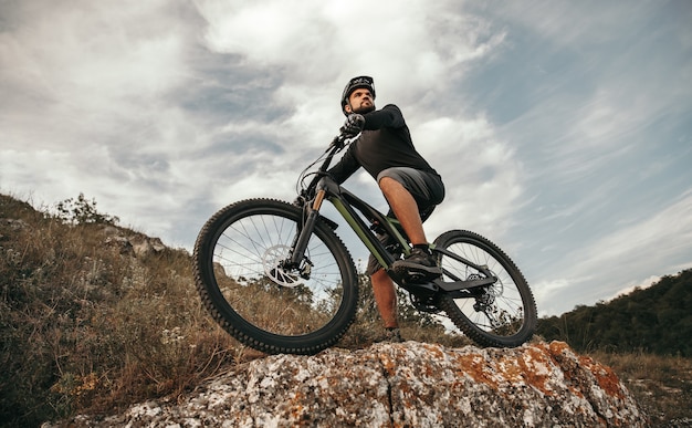 電子マウンテンバイクに乗って、曇り空に対して石の丘から環境を観察するスポーティーな男性自転車のローアングル
