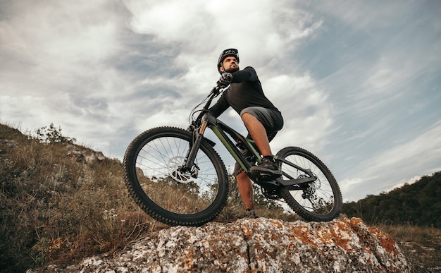 Low angle of sportive male bicyclist riding electronic mountain bike and observing environment from stony hill against cloudy sky