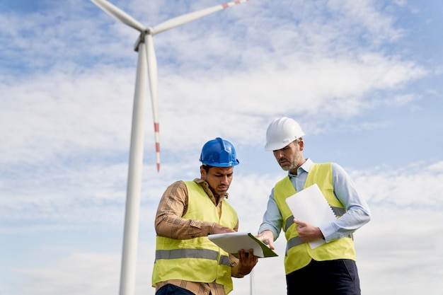 Low angle of two engineers standing on wind turbine field and discussing over documents