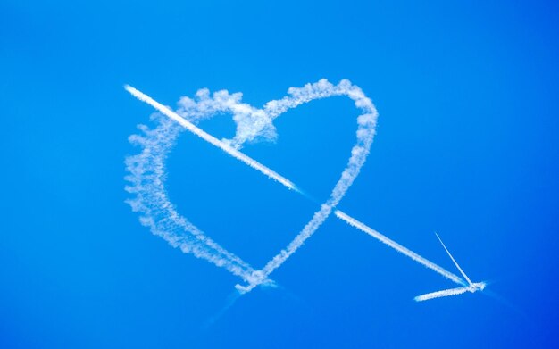 Low angle view of arrow and heart vapor trail in blue sky