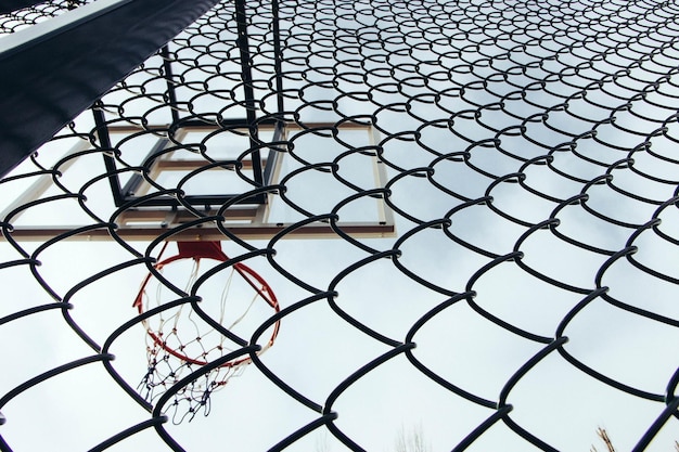 Foto vista a basso angolo del cerchio da basket vista attraverso una recinzione a catena contro un cielo limpido