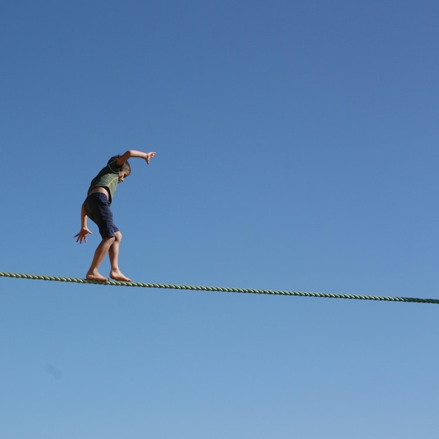 Low angle view of boy walking on tightrope against clear blue sky