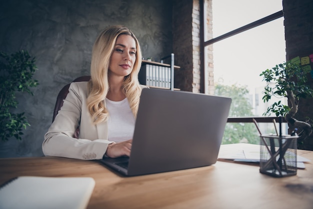 Low below angle view  cheerful beautiful blonde haired entrepreneur sitting at desktop with laptop and notepad