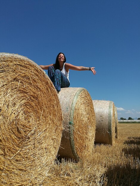 Photo low angle view of cheerful young woman sitting on hay bale against clear sky