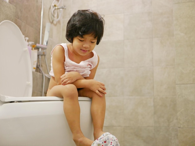 Low angle view of cute girl sitting on toilet bowl in bathroom