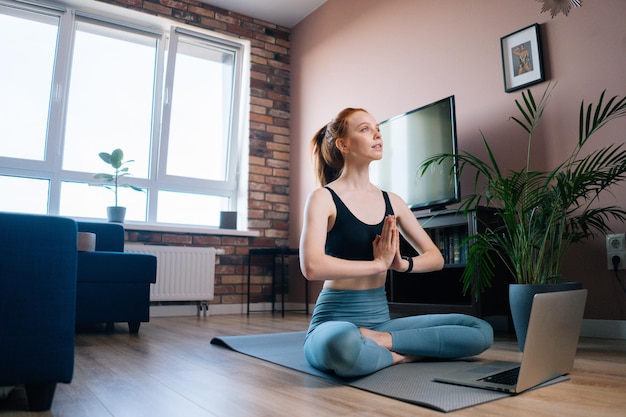 Low-angle view of happy redhead young woman wearing sportswear is meditating in lotus position sitting on yoga mat near laptop. Peaceful red-haired lady holding hands in Namaste pose near chest.