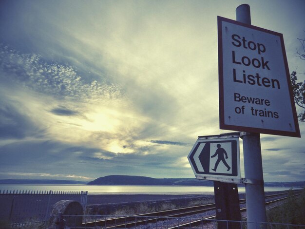 Photo low angle view of information sign by railroad tracks and sea against cloudy sky