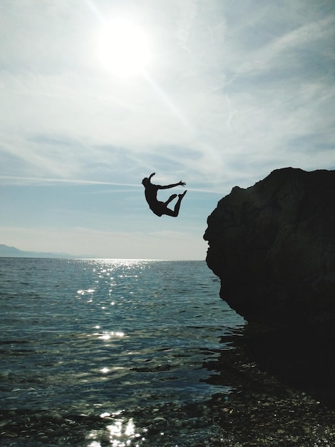 Foto vista a bassa angolazione di un uomo che salta da una scogliera sul mare contro il cielo