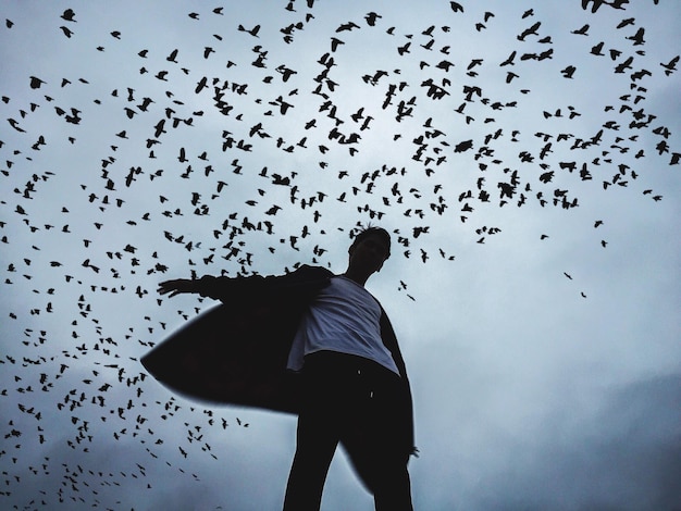 low angle view of man standing under silhouette birds flying against sky
