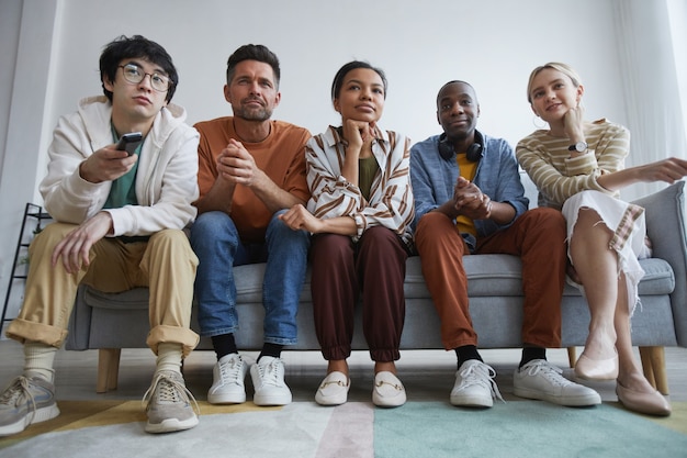 Foto inquadratura dal basso in un gruppo multietnico di amici che guardano la tv a casa insieme seduti in fila sul divano