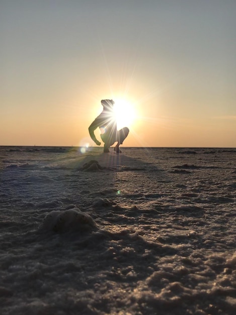 Low angle view of person crouching on snow covered land against sky during sunset