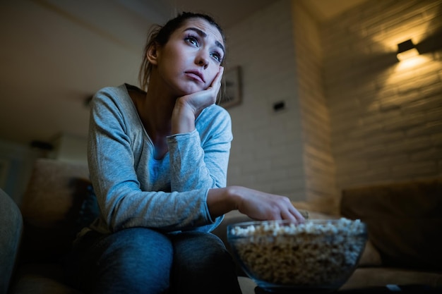 Low angle view of sad woman watching TV and eating popcorn in the evening at home