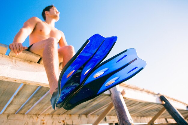 Foto vista ad angolo basso di un uomo senza camicia che indossa pinne da immersione mentre è seduto sul molo contro un cielo blu limpido