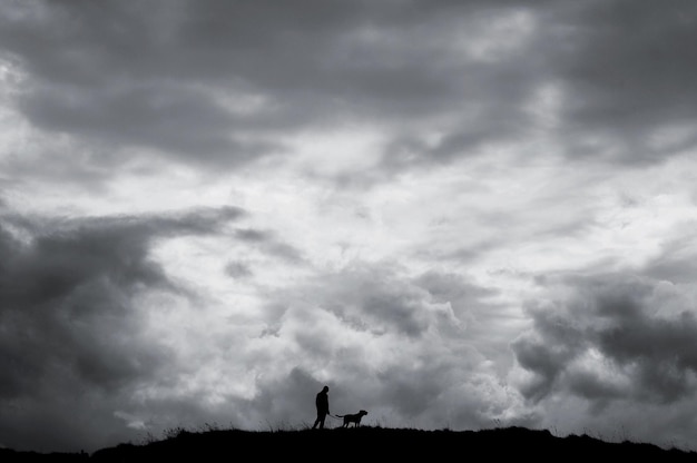 low angle view of silhouette man with dog walking on field against cloudy sky during sunset