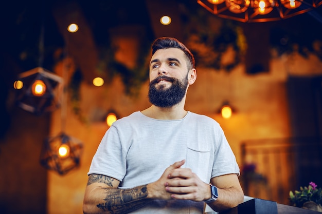 Low angle view of smiling caucasian bearded tattooed hipster leaning on table in cafe and looking away.