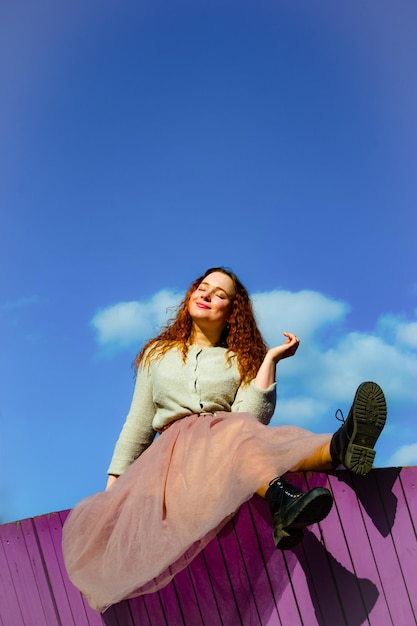 Low angle view of smiling young woman sitting on retaining wall against sky