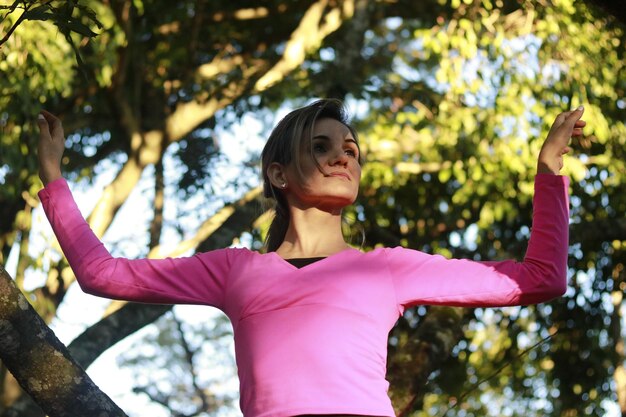 Foto vista ad angolo basso di una donna che guarda via con le braccia alzate mentre è in piedi contro gli alberi