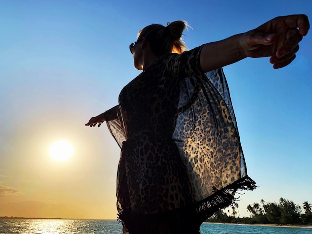 Photo low angle view of woman with arms outstretched standing by sea against sky during sunset