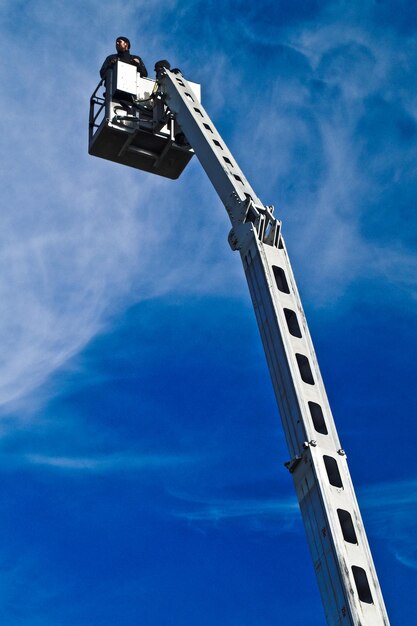 Low angle view of worker standing in crane against blue sky on sunny day