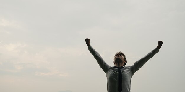 Low angle view of young businessman standing with his arms raised under a cloudy sky.