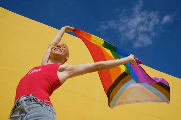Low angle view of a young non-binary person holding and raising up a rainbow flag while posing outdoors with blue sky on background. Equality, rights and identity gender concept.