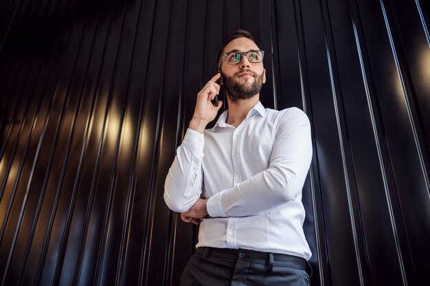 Low angle view of young pensive geeky man leaning on the wall and calling boss.