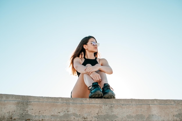 明るい空の前で壁に寄りかかって座っている若い女性の低角度の写真