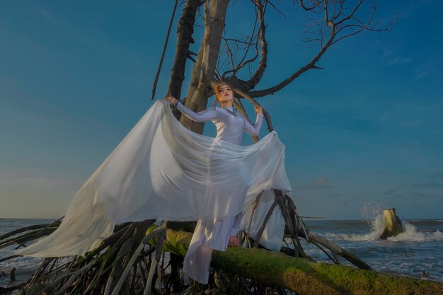 Photo low angle view of young woman with scarf posing on bare tree at beach