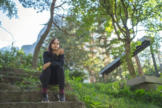 Low angle of young female traveler in activewear sitting on old stone stairs and drinking coffee while resting during walk in green botanical park