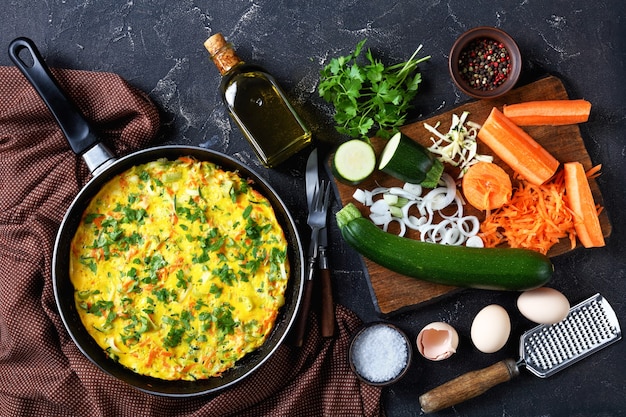 Low-carb zucchini and carrot frittata served on a frying pan on a dark concrete table with a bottle of olive oil, parsley, cutlery, peppercorns, and fresh vegetables, top view, flat lay, close-up