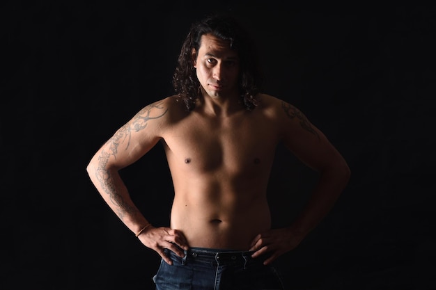 Low key portrait of a man with tattoo looking at camera and hands on waist on black background