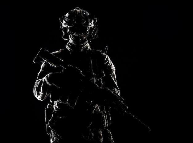 Low key studio portrait of army special forces elite soldier with hidden behind mask and glasses face, battle helmet, tactical radio headset, standing with assault rifle equipped silencer in darkness