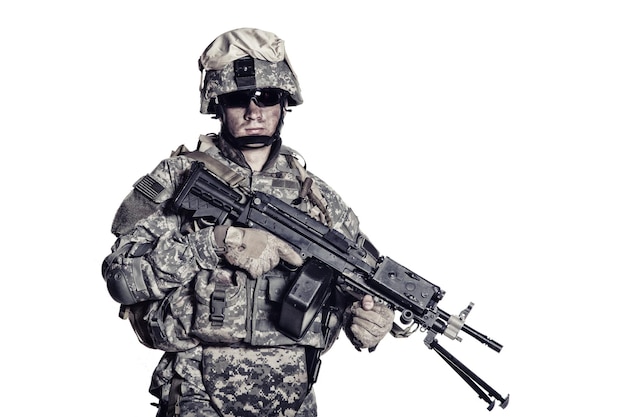 Low key studio portrait of US Armed Forces soldier in protective camouflage, helmet and tactical sunglasses holding light machine gun and looking in camera desaturated, isolated on white background