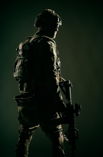 Low key studio shoot of army special forces soldier, commando fighter in mask, ballistic glasses, tactical helmet and battle uniform, holding short barrel service rifle, looking back over shoulder