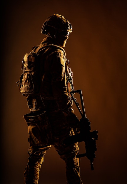 Low key studio shoot of army special forces soldier, commando fighter in mask, ballistic glasses, tactical helmet and battle uniform, holding short barrel service rifle, looking back over shoulder